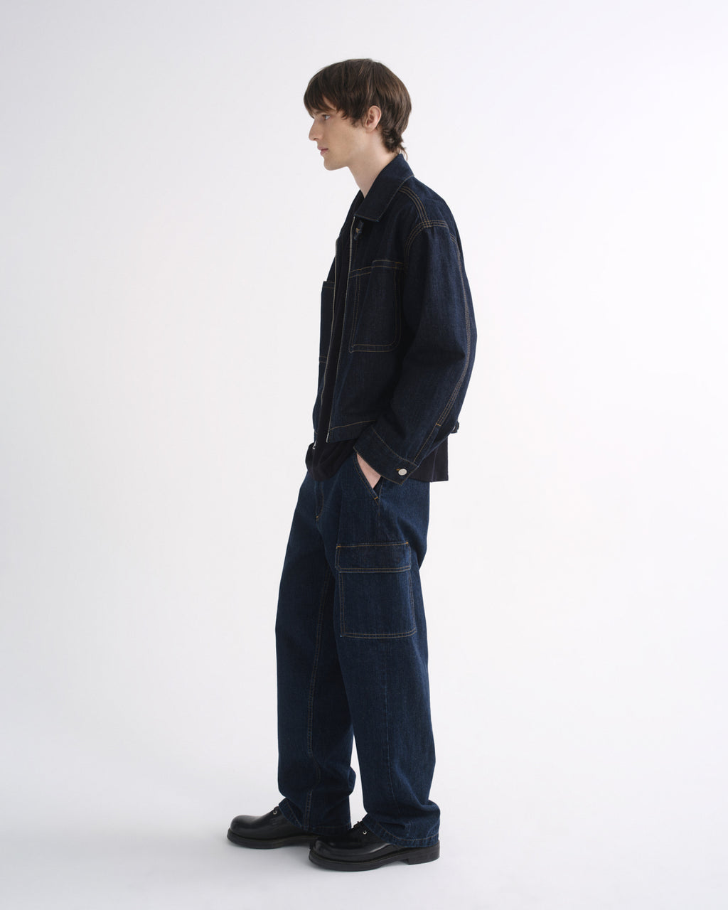 Person wearing a dark indigo denim jacket with matching dark indigo pants and black shoes, side view, on a grey background