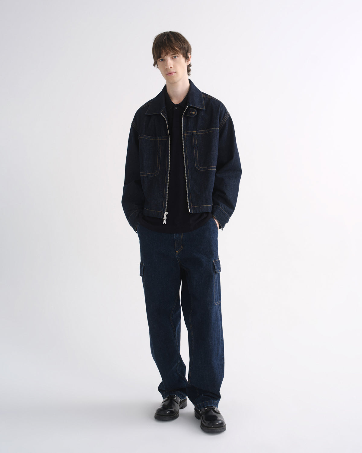 Person wearing a dark indigo denim jacket with matching dark indigo pants and black shoes on a grey background