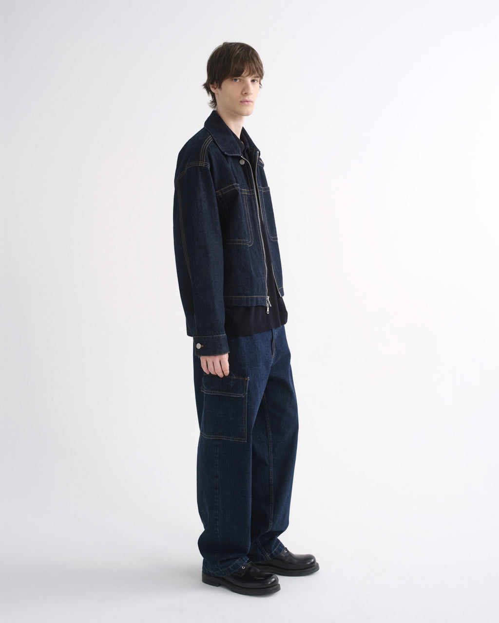 Person wearing a dark indigo denim jacket with matching dark indigo pants and black shoes, side view, on a grey background