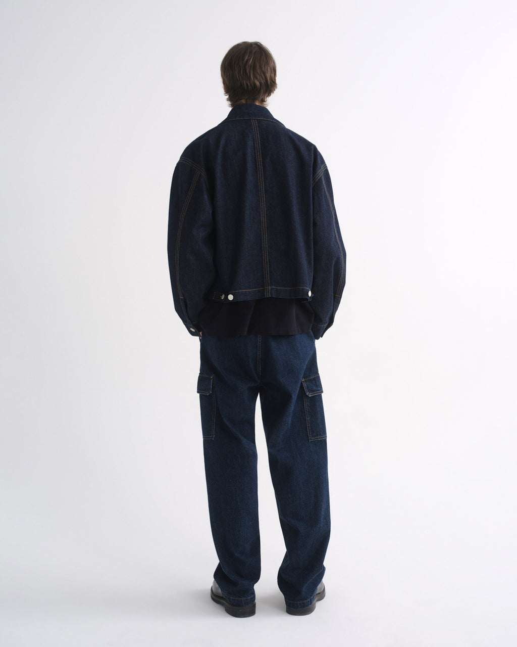 Person wearing a dark indigo denim jacket with matching dark indigo pants and black shoes, back view, on a grey background