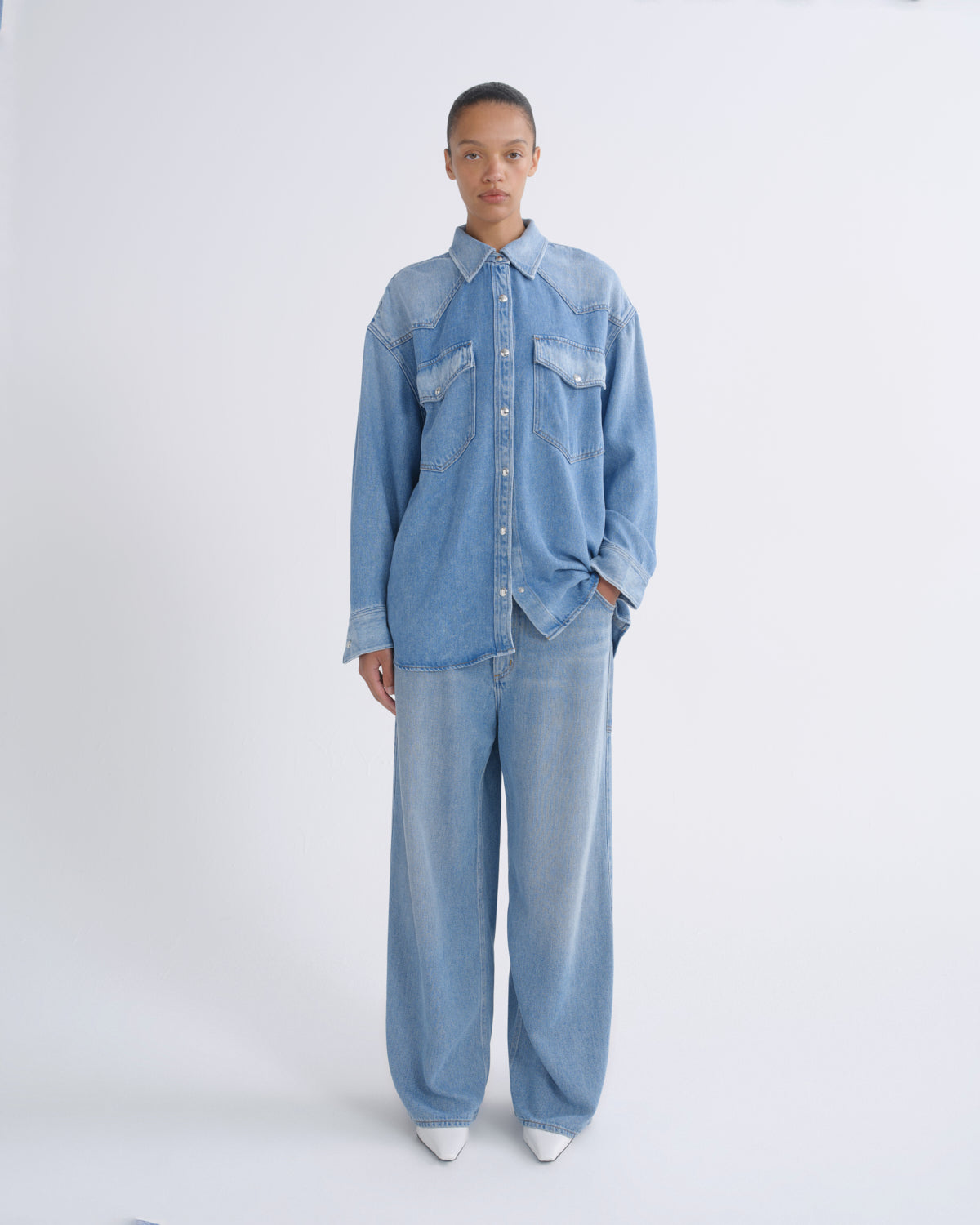 A model stands against a plain white studio background wearing a light blue denim button-up shirt and matching wide-leg denim pants.