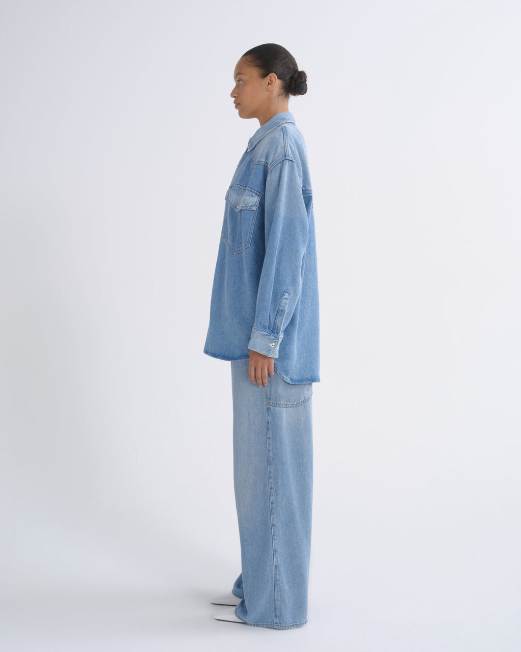 A model shown in profile wearing a light blue denim shirt and matching wide-leg denim pants against a plain white studio background.