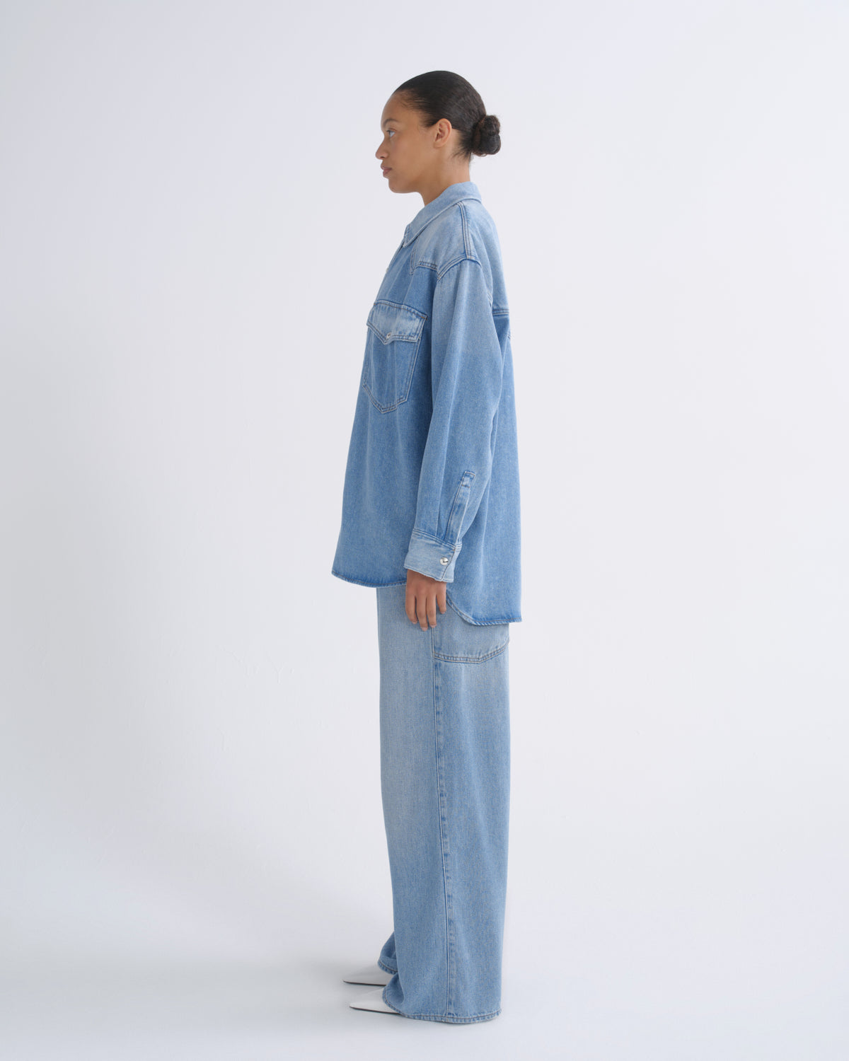 A model shown in profile wearing a light blue denim shirt and matching wide-leg denim pants against a plain white studio background.
