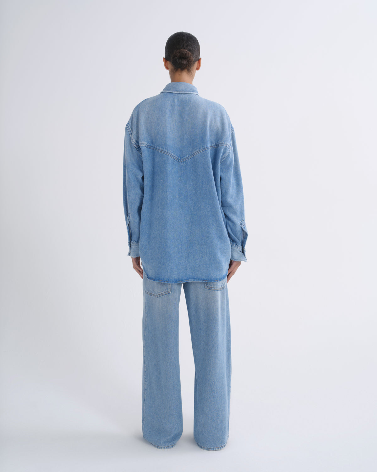 A model viewed from the back wearing a light blue denim shirt and matching wide-leg jeans against a plain white studio background.