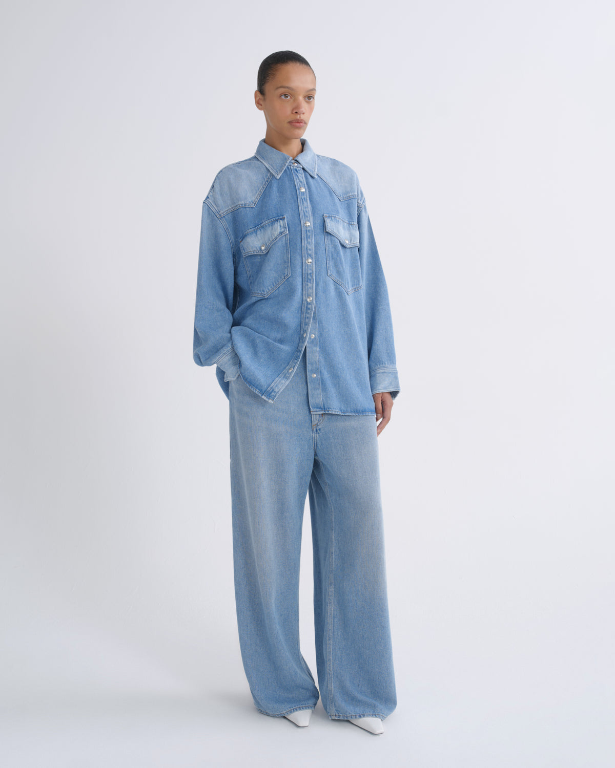 A model stands against a light gray studio background wearing a light blue denim shirt and matching wide-leg jeans.