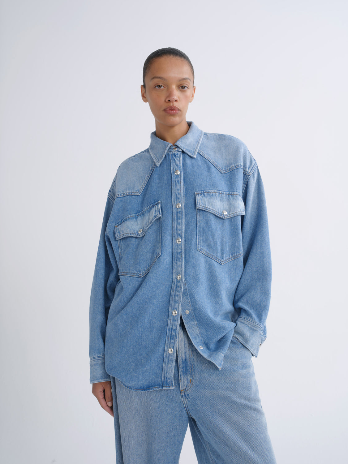 A model wearing a light blue denim long-sleeve button-up shirt with western-style chest pockets, paired with matching light blue denim pants, against a plain light gray studio background.