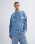 A model wearing a light blue denim western-style shirt with chest pockets and matching light blue denim jeans, standing against a plain light gray studio background.