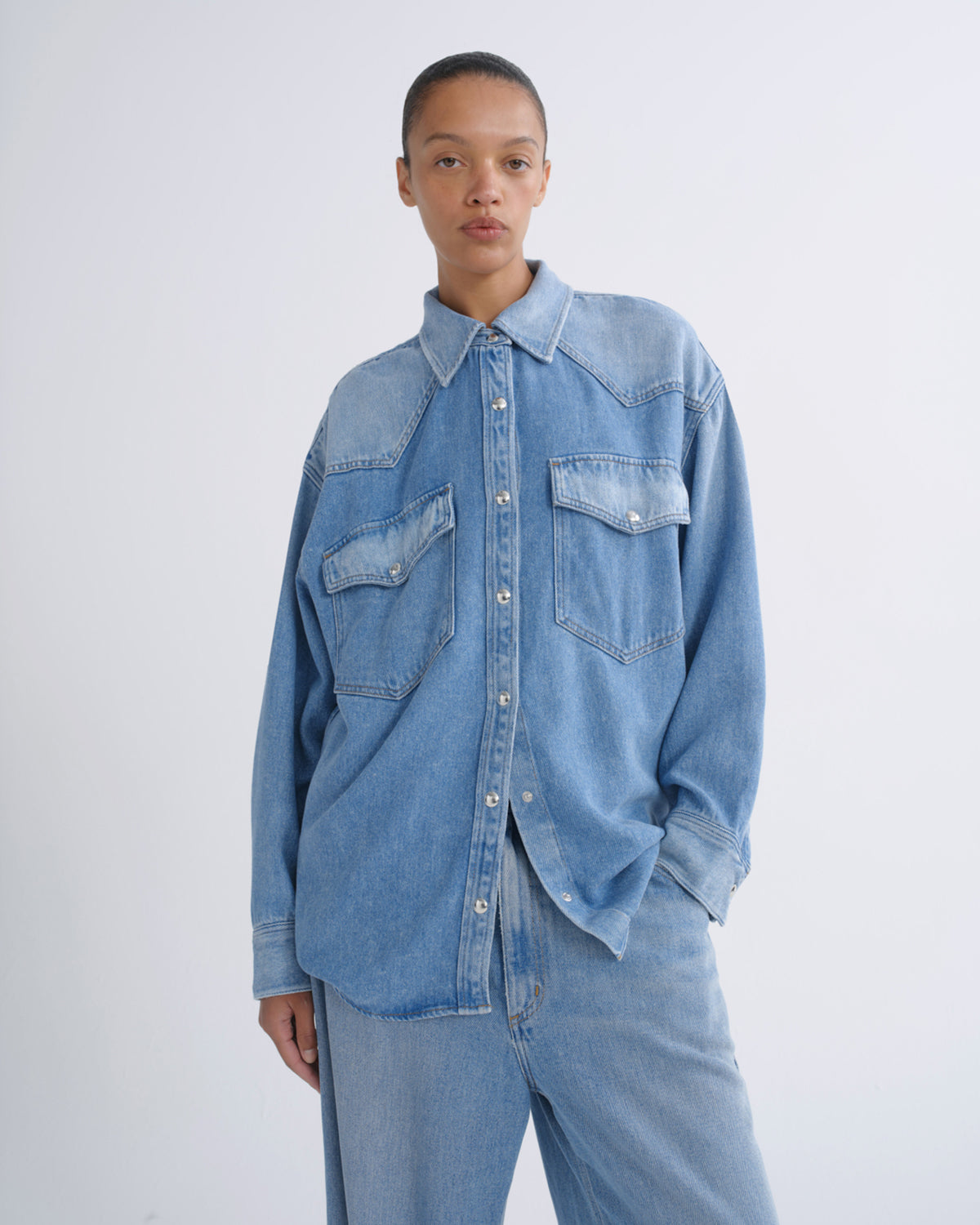 A model wearing a light blue denim western-style shirt with chest pockets and matching light blue denim jeans, standing against a plain light gray studio background.
