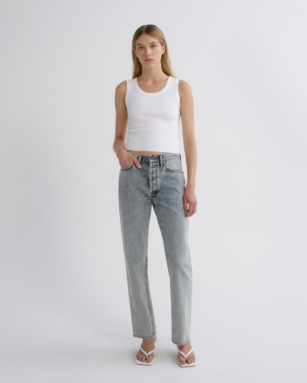 A model stands against a light gray studio background wearing a white ribbed tank top, light-wash straight-leg denim jeans, and white heeled sandals.