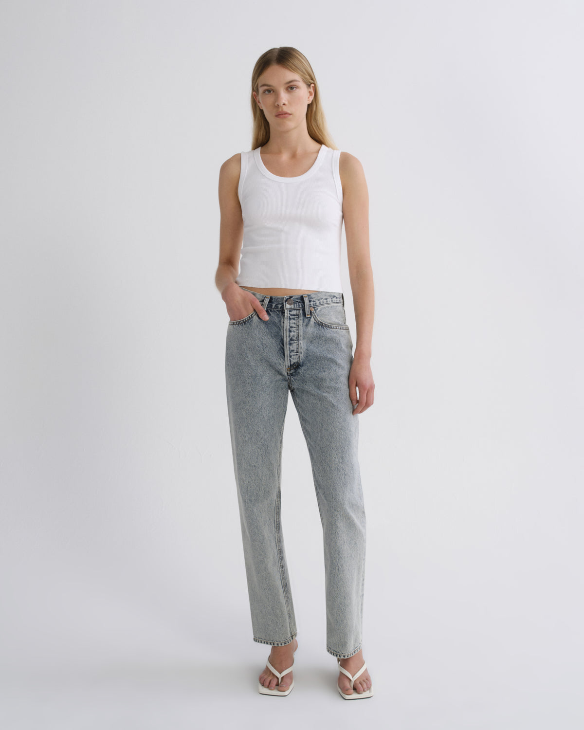 A model stands against a light gray studio background wearing a white ribbed tank top, light-wash straight-leg denim jeans, and white heeled sandals.