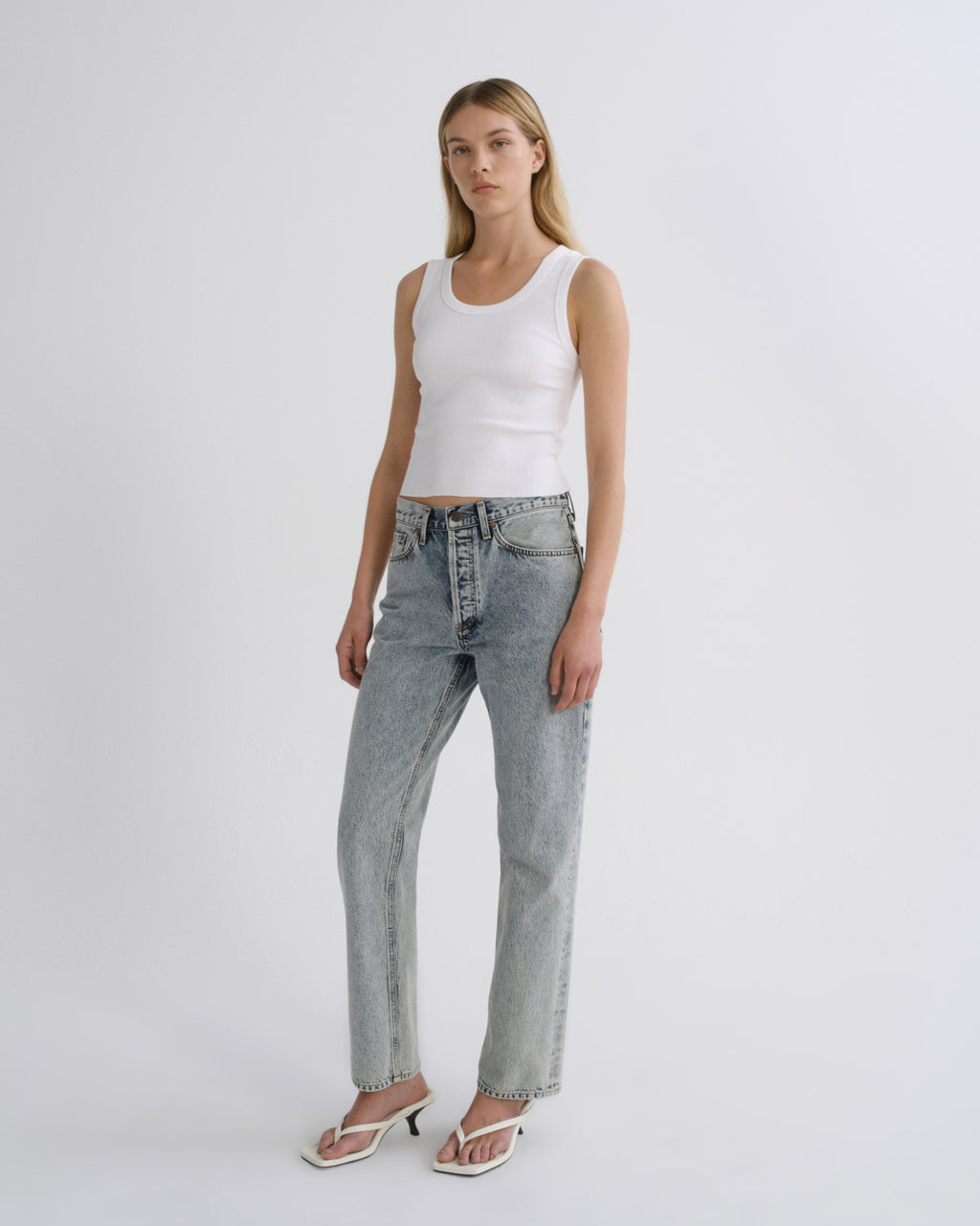 A woman modeling a white ribbed tank top and light-wash blue straight-leg jeans with white heeled sandals, standing against a plain light gray studio background.