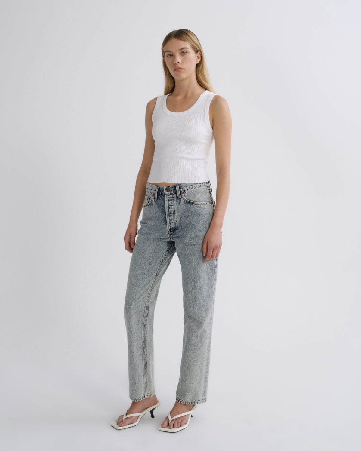 A woman modeling a white ribbed tank top and light-wash blue straight-leg jeans with white heeled sandals, standing against a plain light gray studio background.