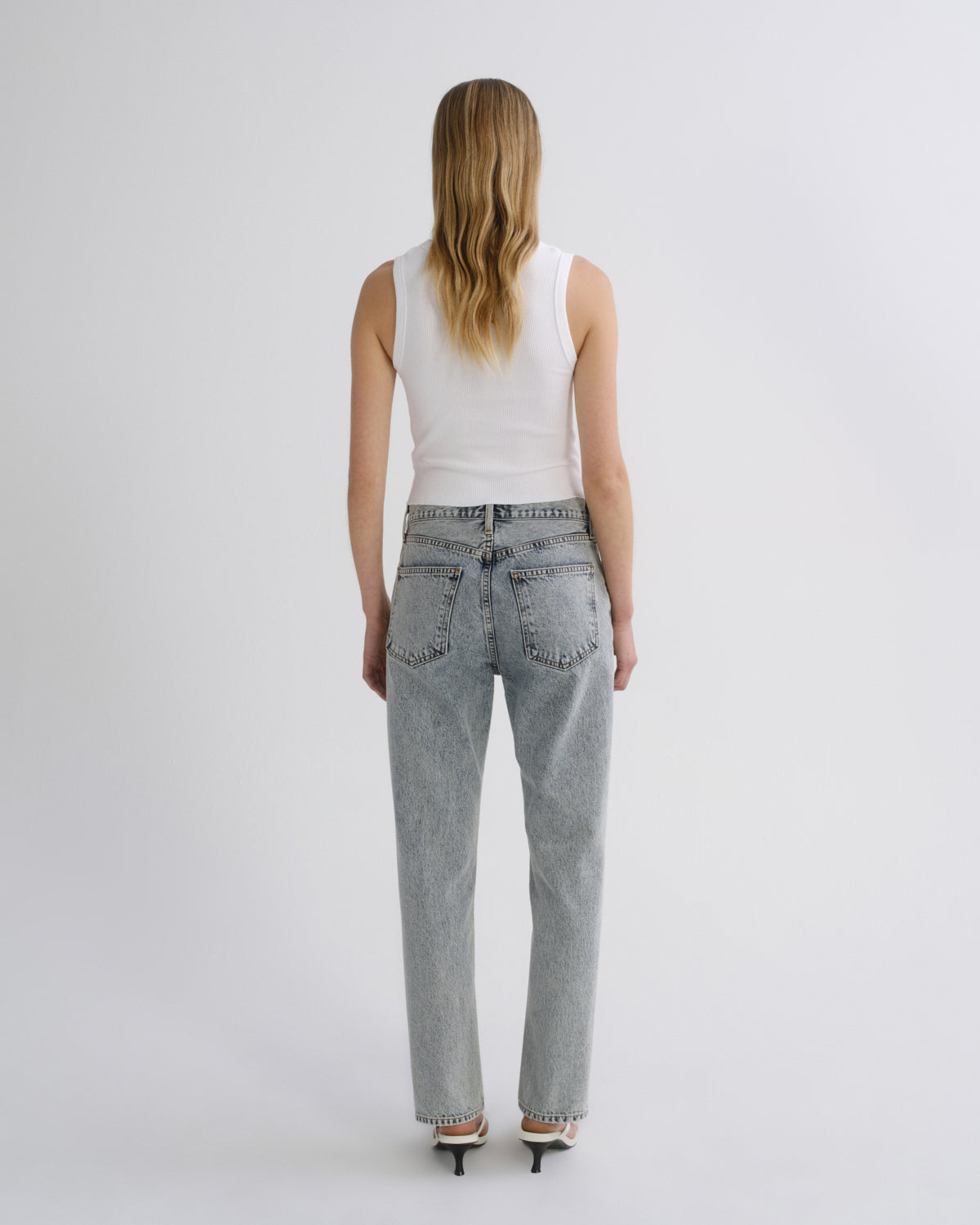 A woman viewed from behind wearing light-wash straight-leg jeans, a white sleeveless top, and black high heels against a plain light gray studio background.