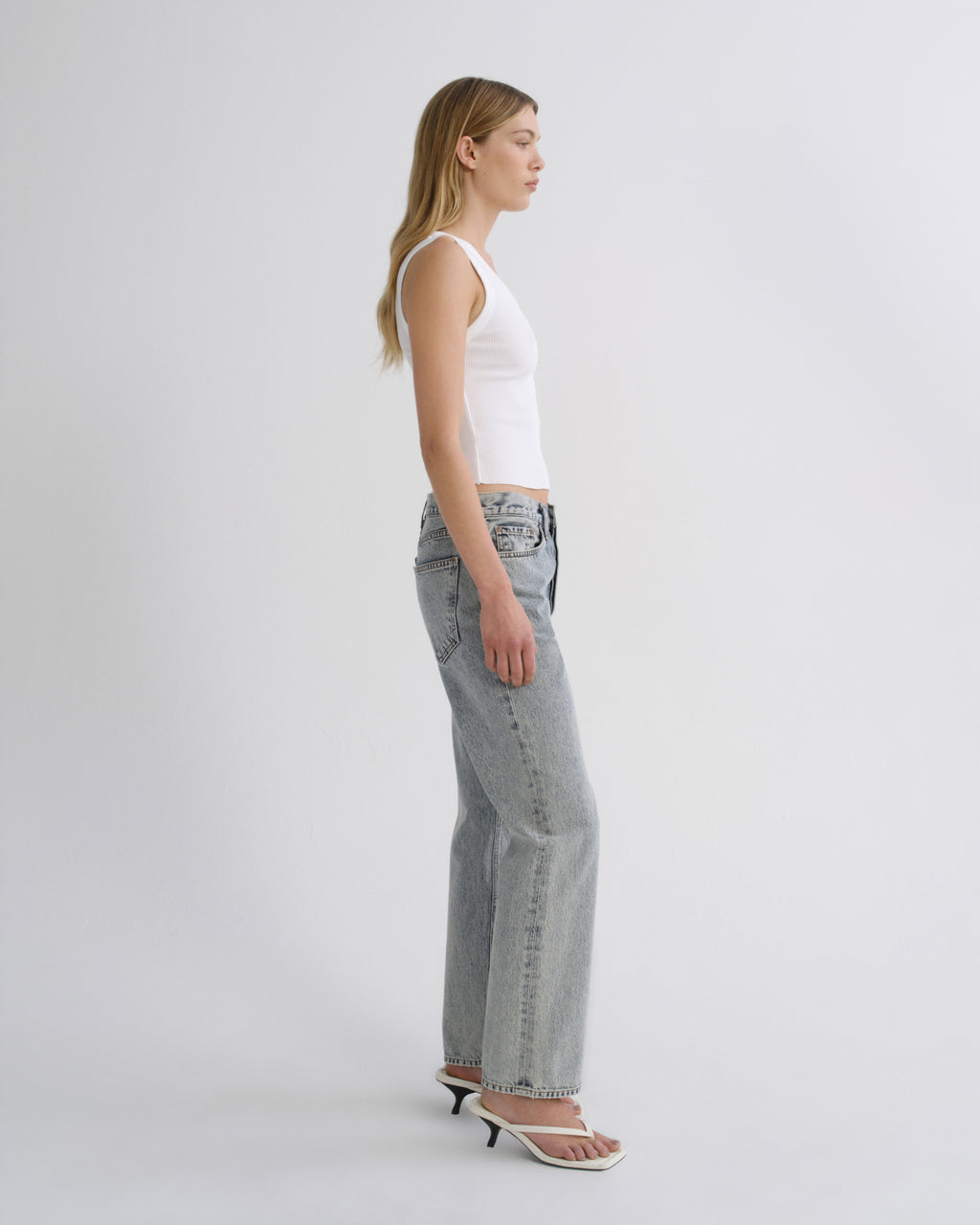 A woman in side profile wearing light-wash wide-leg jeans, a white ribbed tank top, and white heeled sandals, standing against a plain light gray studio background.