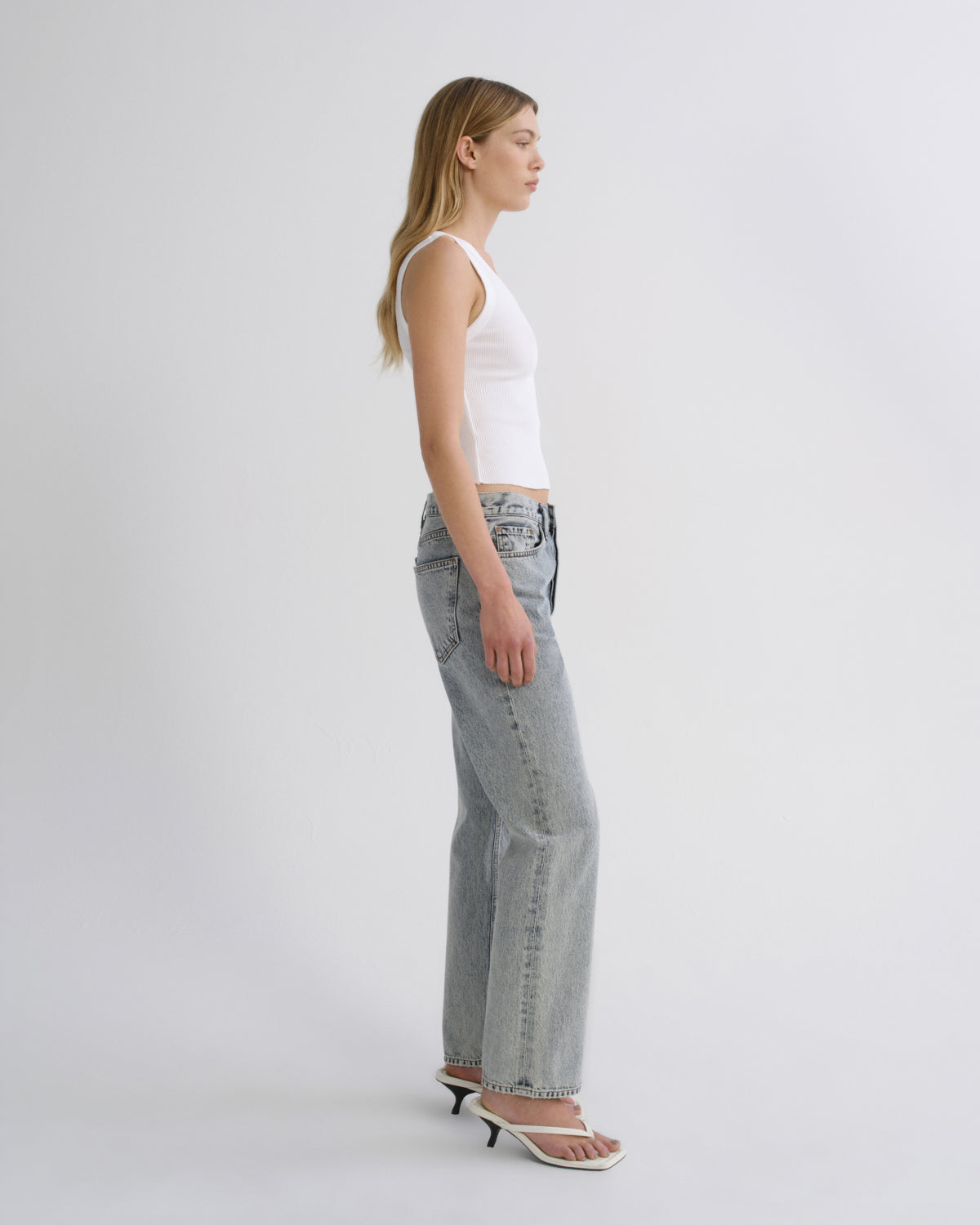 A woman in side profile wearing light-wash wide-leg jeans, a white ribbed tank top, and white heeled sandals, standing against a plain light gray studio background.