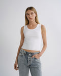 A woman modeling a white ribbed crop tank top paired with light-wash blue jeans against a plain light gray studio background.