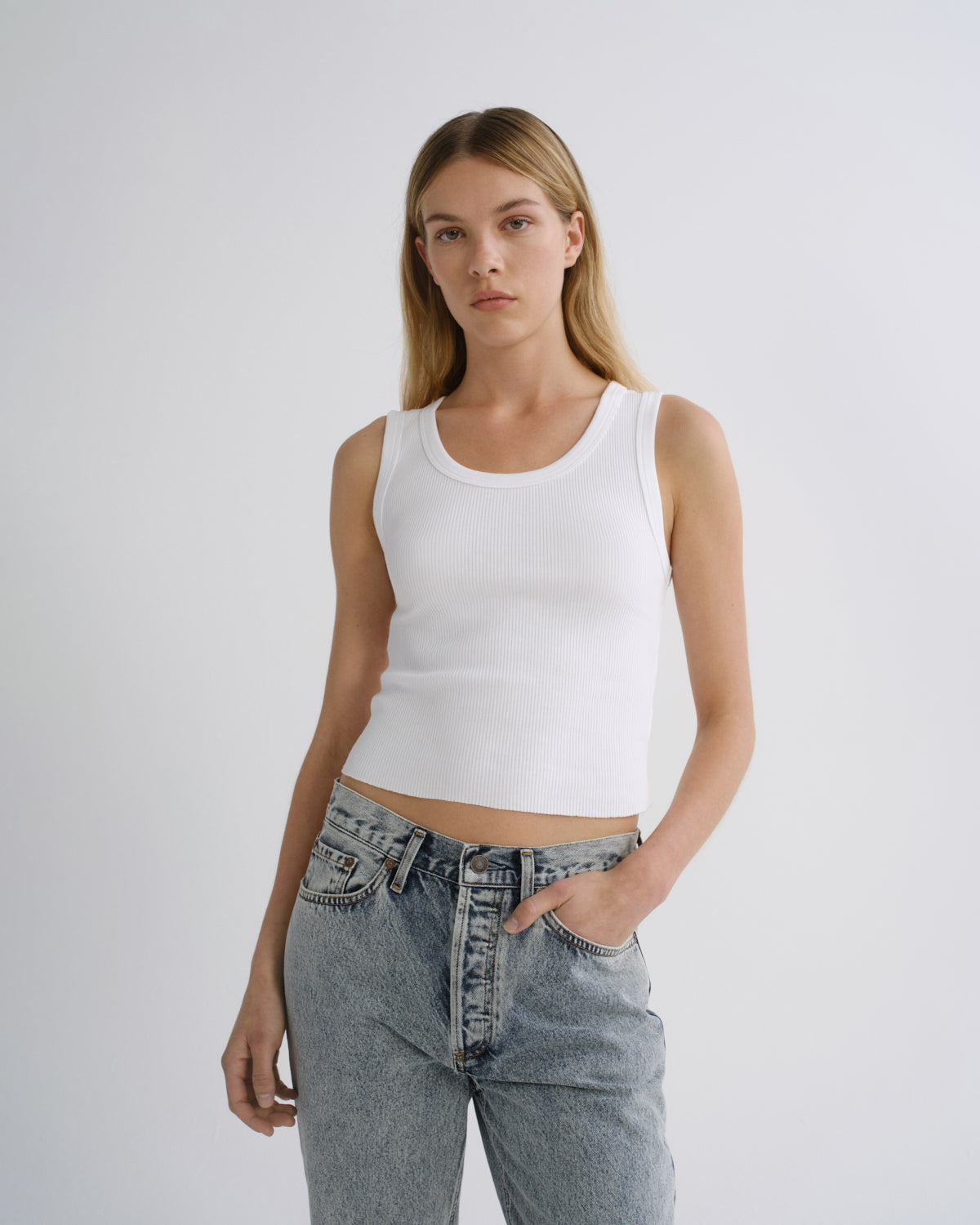 A woman modeling a white ribbed crop tank top paired with light-wash blue jeans against a plain light gray studio background.