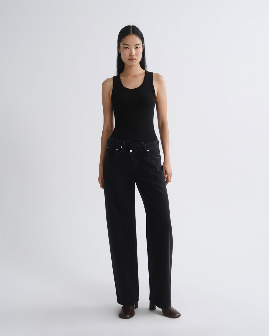 A model stands against a light gray studio background wearing a black sleeveless top and black wide-leg jeans, paired with dark brown shoes.
