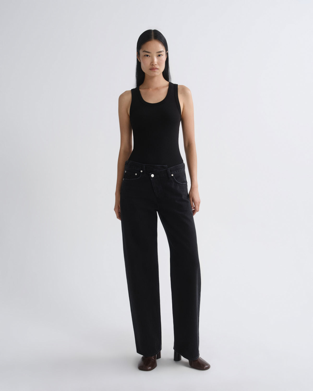 A model stands against a light gray studio background wearing a black sleeveless top and black wide-leg jeans, paired with dark brown shoes.
