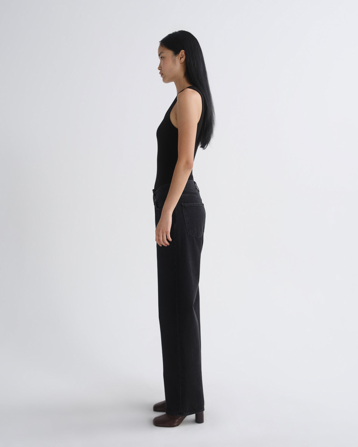 A woman stands in profile wearing a black sleeveless top and black wide-leg pants against a plain light gray studio background.