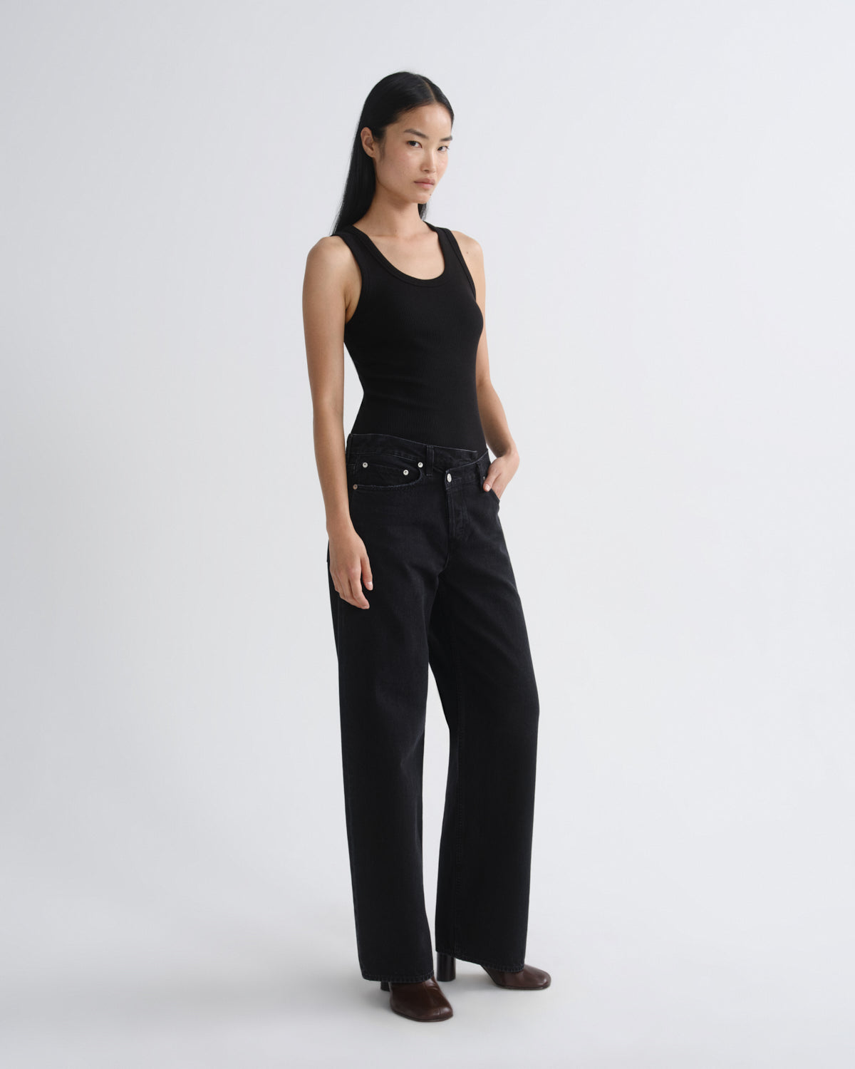 A woman modeling a black sleeveless tank top and black wide-leg jeans against a plain light gray studio background.