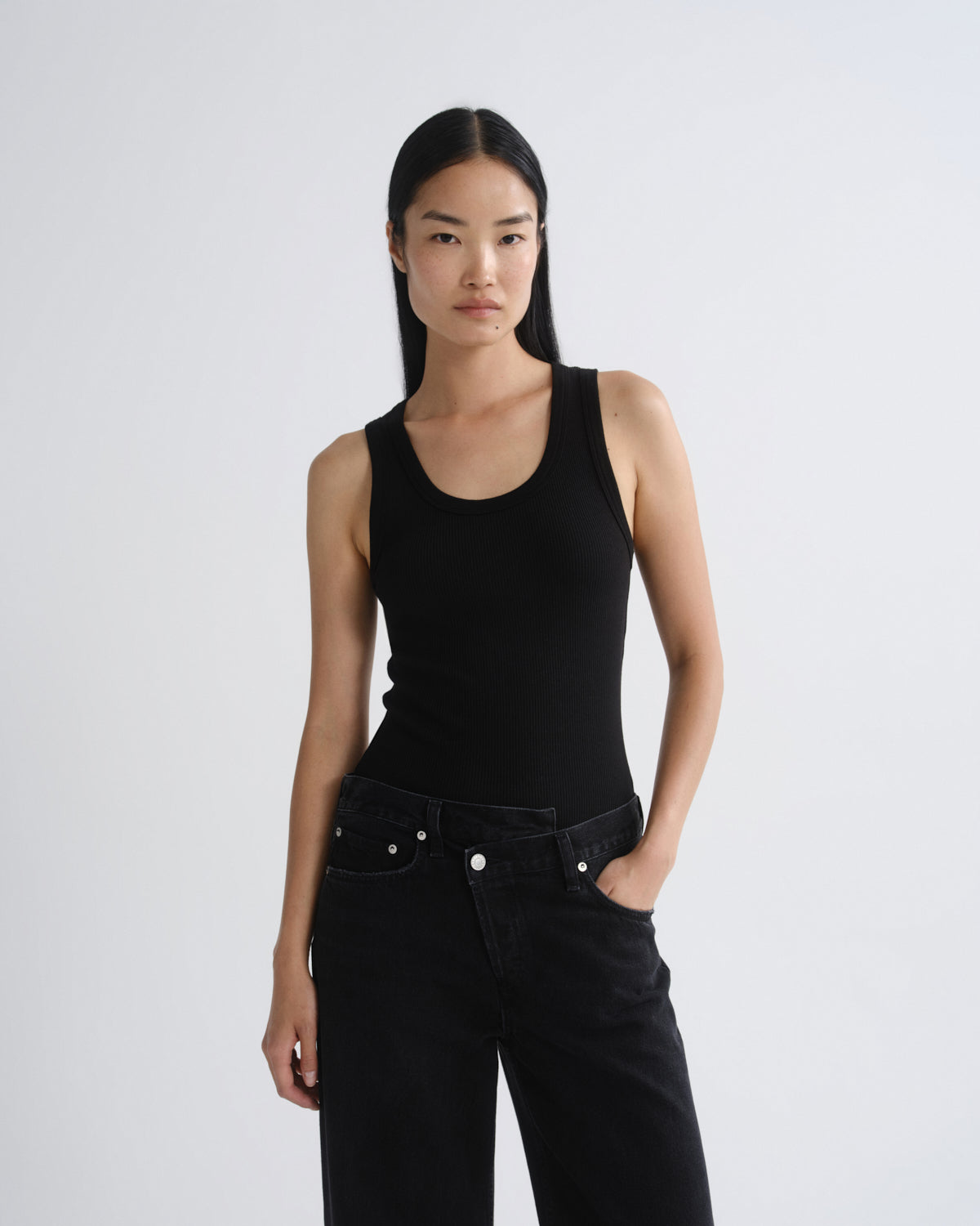 A model wearing a black ribbed scoop-neck tank top and black jeans, standing against a light gray studio background.