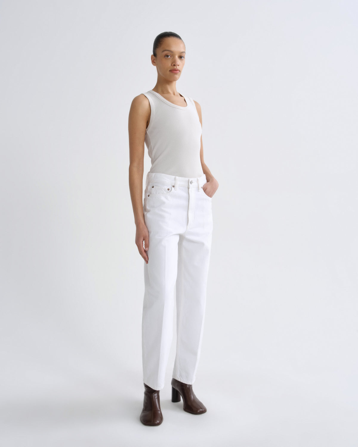 A model stands against a light gray studio background wearing a white ribbed tank top, white wide-leg jeans, and dark brown ankle boots.