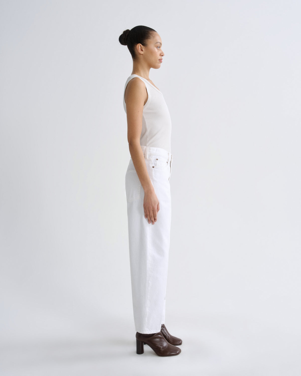 A model in profile wearing a white sleeveless top and white wide-leg jeans, paired with dark brown heeled ankle boots, against a plain light gray studio background.