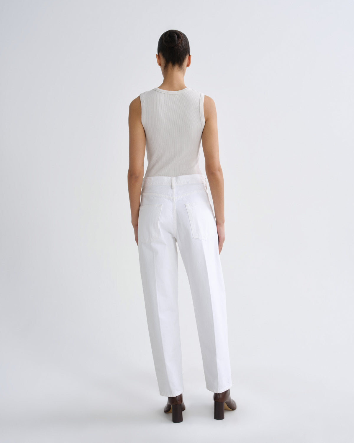 A model viewed from the back wearing a sleeveless light beige top and white trousers, standing against a plain light gray studio background.