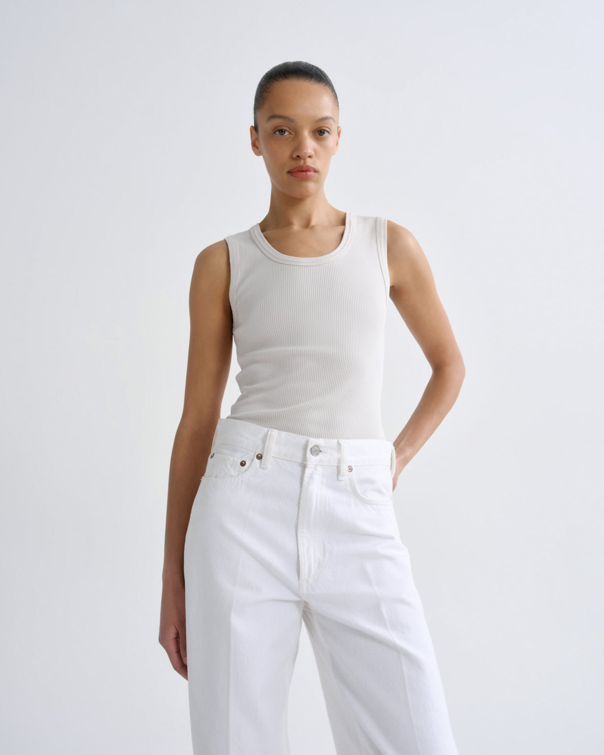 A model wearing a fitted white ribbed sleeveless tank top and white high-waisted wide-leg jeans, standing against a light gray studio background.