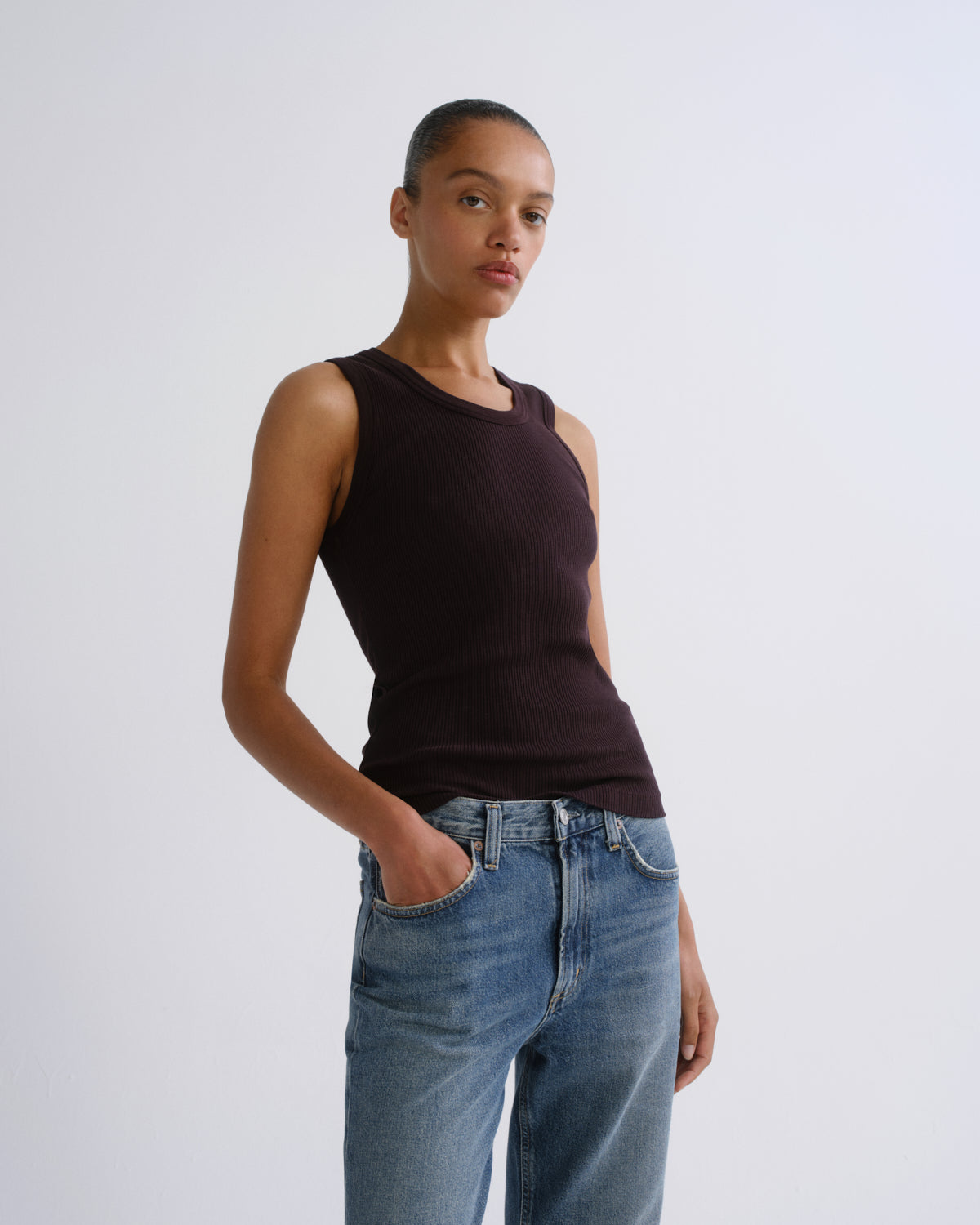 A model wearing a dark brown ribbed sleeveless top and blue jeans, standing against a light gray studio background.