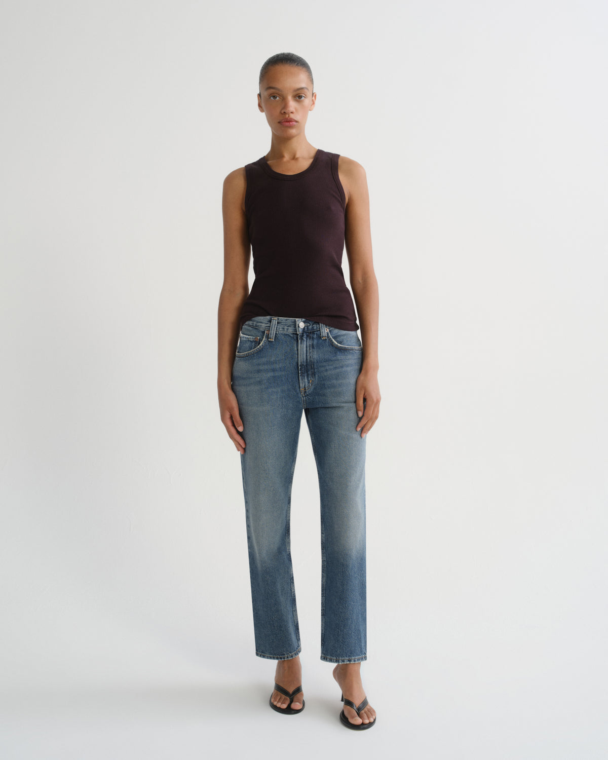 A model stands against a light gray studio background wearing a sleeveless dark plum top, blue cropped jeans with a faded wash, and black sandals.