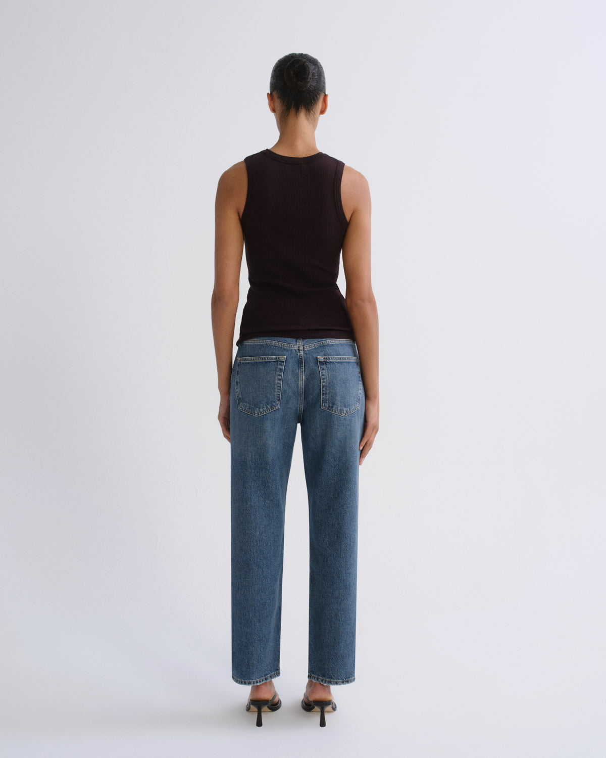 A model shown from the back wearing a dark brown sleeveless top, blue wide-leg jeans, and black heels, set against a plain light gray studio background.