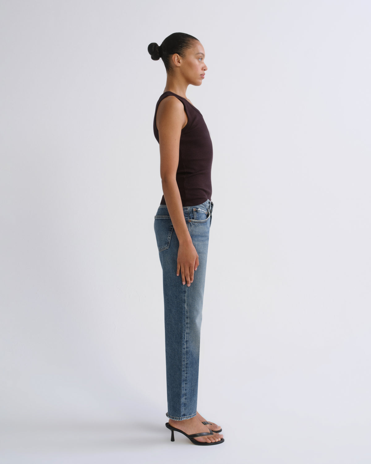 A woman in profile wearing a dark brown sleeveless top, light-wash blue jeans, and black heeled sandals, standing against a plain light gray studio background.