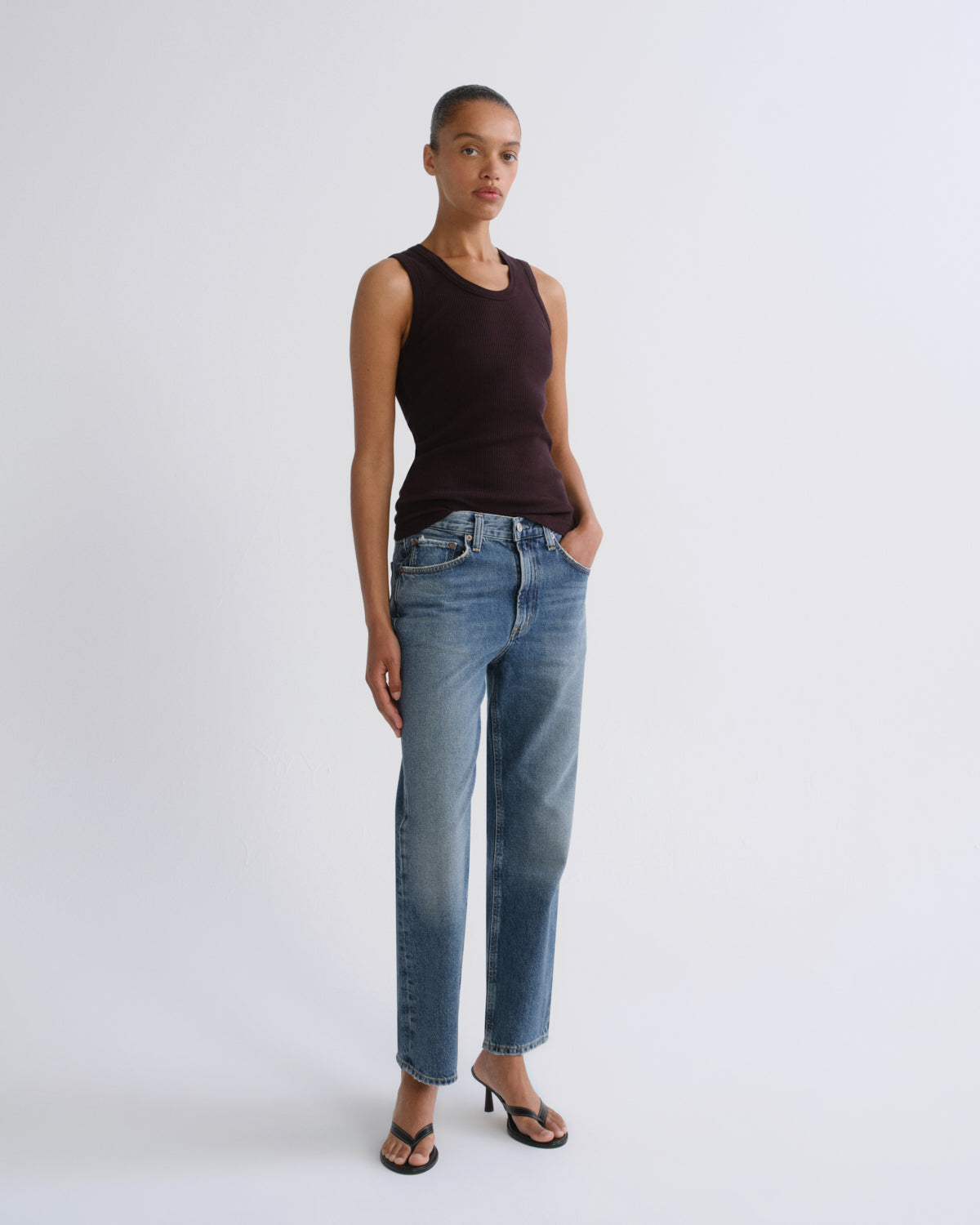 A model stands against a light gray studio background wearing a dark brown sleeveless top, light-wash blue straight-leg jeans, and black heeled sandals.