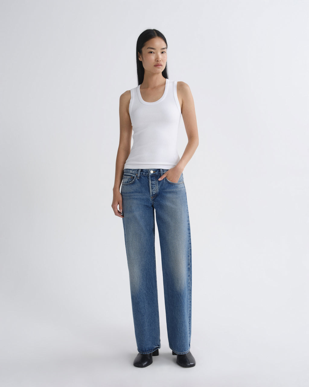 A model stands against a light gray studio background wearing a white ribbed tank top, wide-leg blue jeans, and black shoes.
