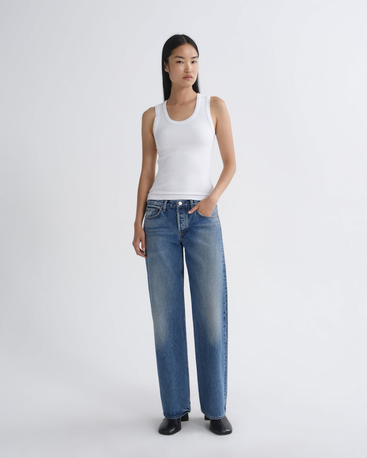 A model stands against a light gray studio background wearing a white ribbed tank top, wide-leg blue jeans, and black shoes.