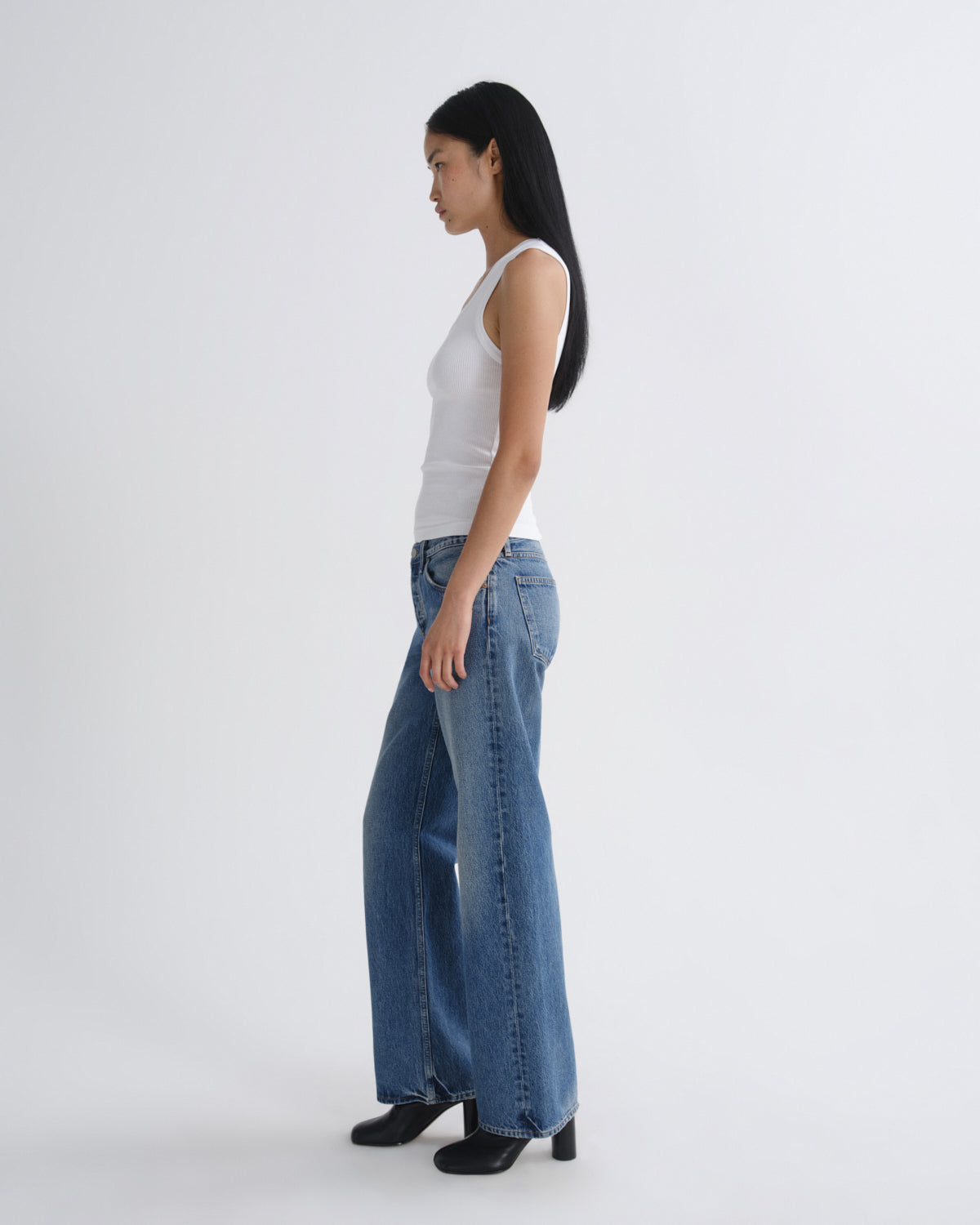 A woman in a white sleeveless top and wide-leg blue jeans stands in profile against a plain light gray studio background.