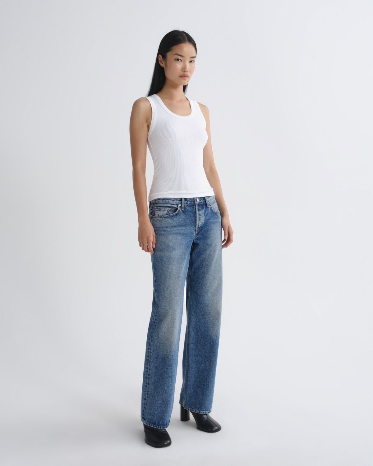 A model wearing a white ribbed tank top and blue wide-leg jeans, standing against a light gray studio background.