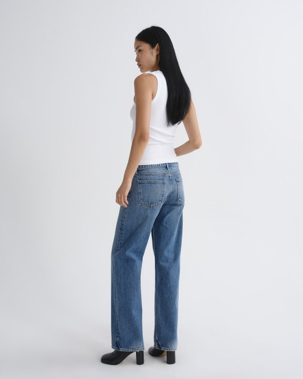 A woman shown from the back wearing a white sleeveless top, blue wide-leg jeans, and black heels, standing against a plain light gray studio background.