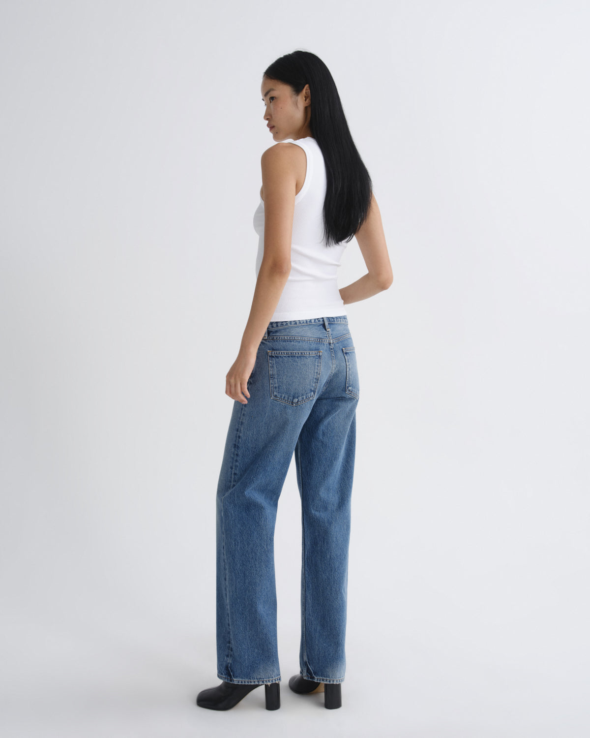 A woman shown from the back wearing a white sleeveless top, blue wide-leg jeans, and black heels, standing against a plain light gray studio background.