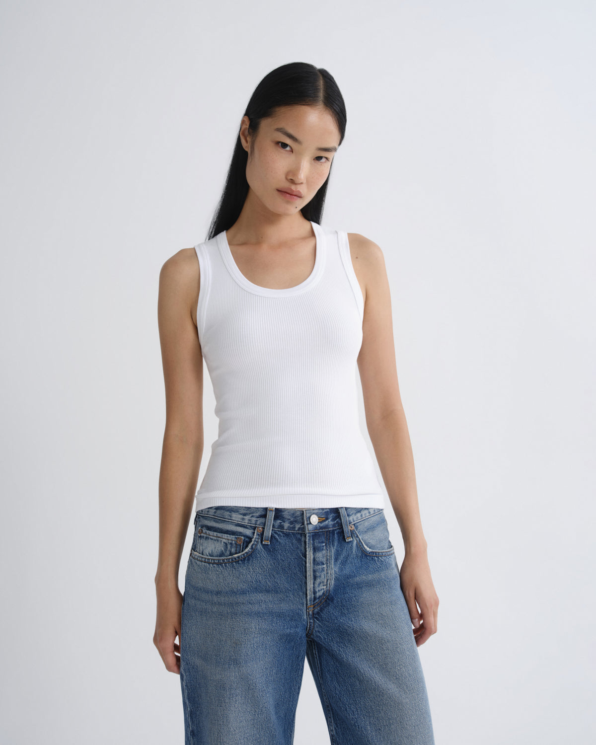 A model wearing a white ribbed tank top and blue jeans, standing against a light gray studio background.