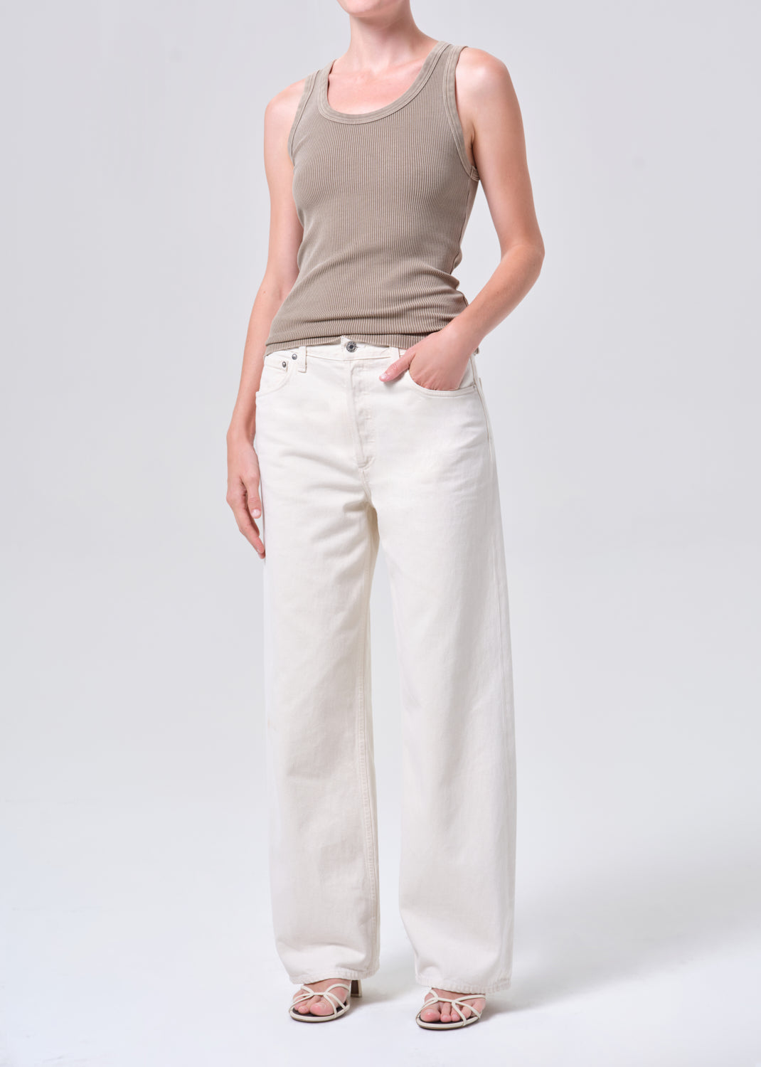 Person wearing a beige tank top and white pants on a light gray background
