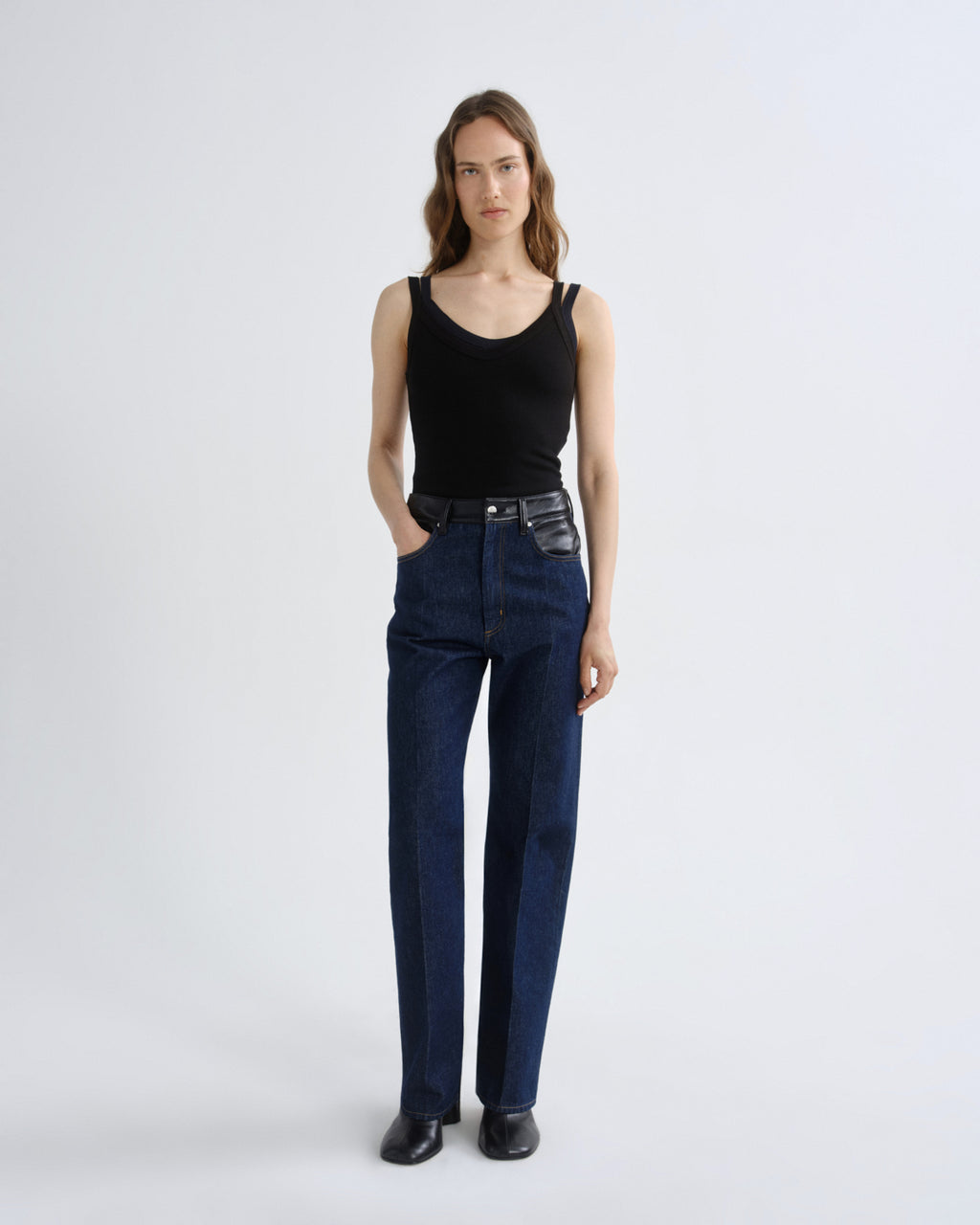 A model stands against a light gray studio background wearing a black ribbed tank top, dark blue straight-leg jeans, and black leather shoes.