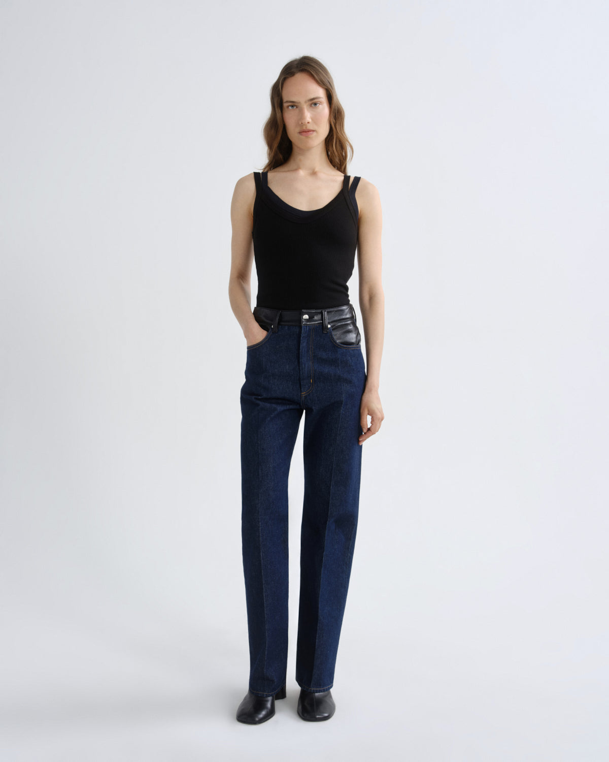 A model stands against a light gray studio background wearing a black ribbed tank top, dark blue straight-leg jeans, and black leather shoes.