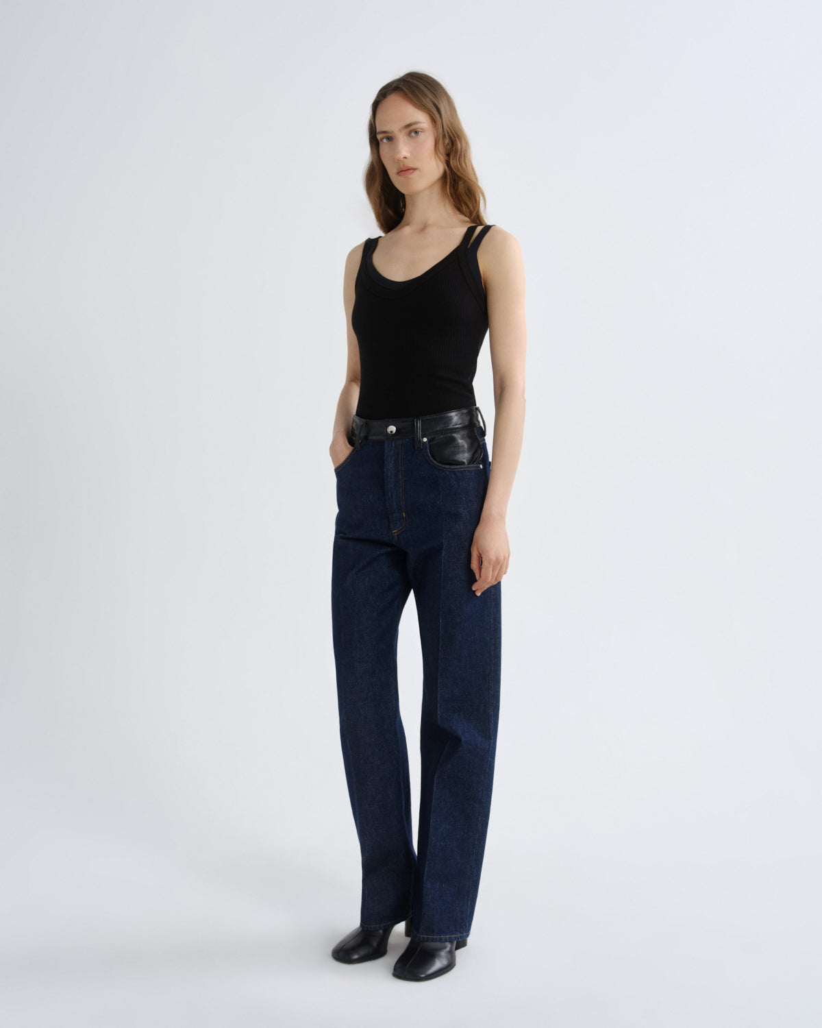 A model stands against a light gray studio background wearing a black ribbed tank top, dark blue straight-leg jeans with a black leather belt, and black leather shoes.