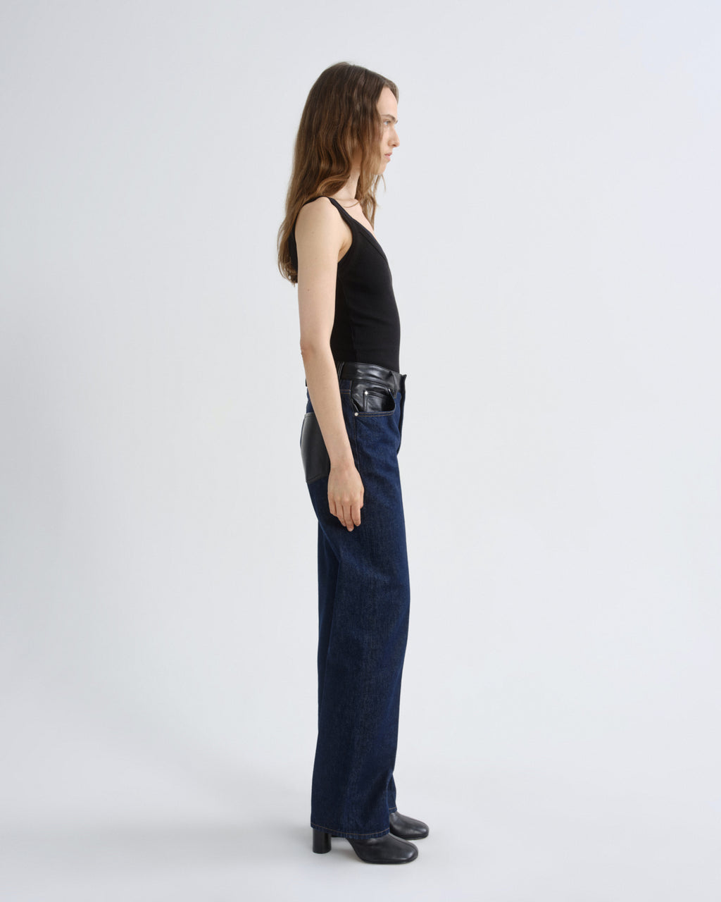 A model shown in profile wearing a black halter top, dark blue wide-leg jeans with black leather side panels, and black shoes, photographed against a light gray studio background.