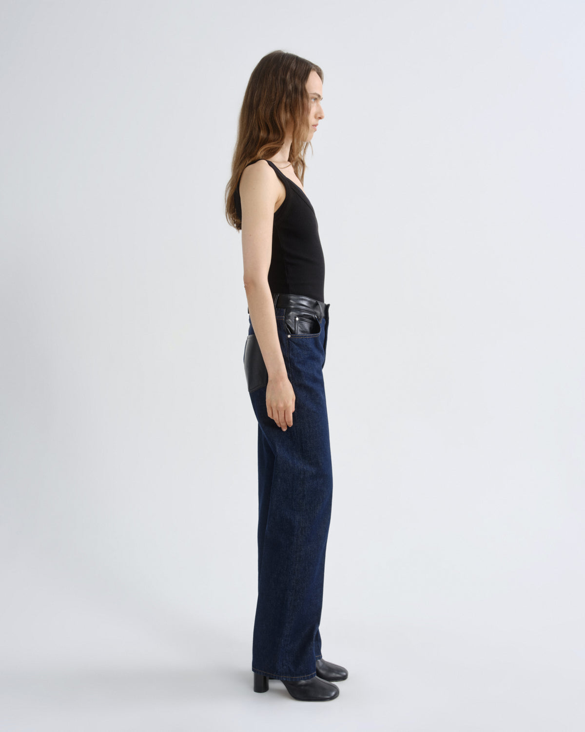 A model shown in profile wearing a black halter top, dark blue wide-leg jeans with black leather side panels, and black shoes, photographed against a light gray studio background.