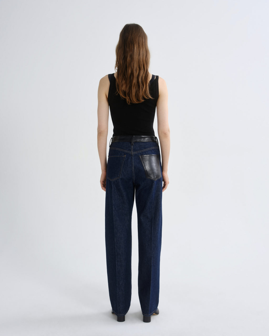 A model viewed from behind wearing dark blue wide-leg jeans with a black leather patch on the back pocket, a black sleeveless top, and black shoes, set against a plain light gray studio background.