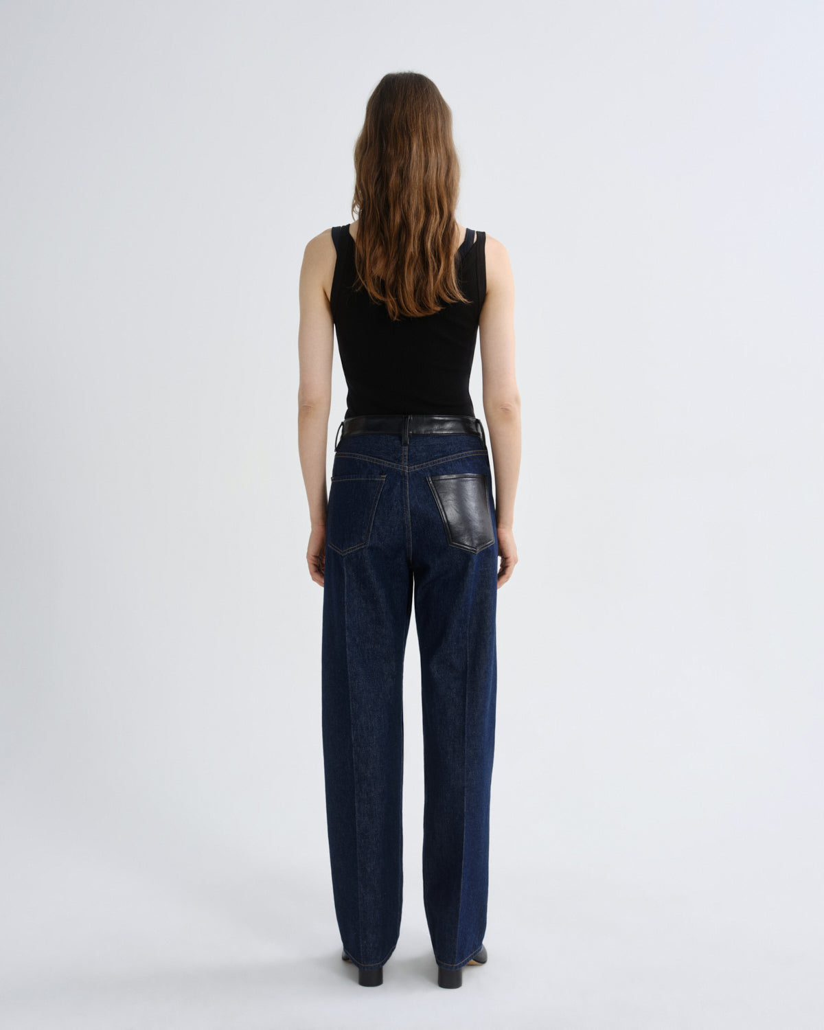 A model viewed from behind wearing dark blue wide-leg jeans with a black leather patch on the back pocket, a black sleeveless top, and black shoes, set against a plain light gray studio background.